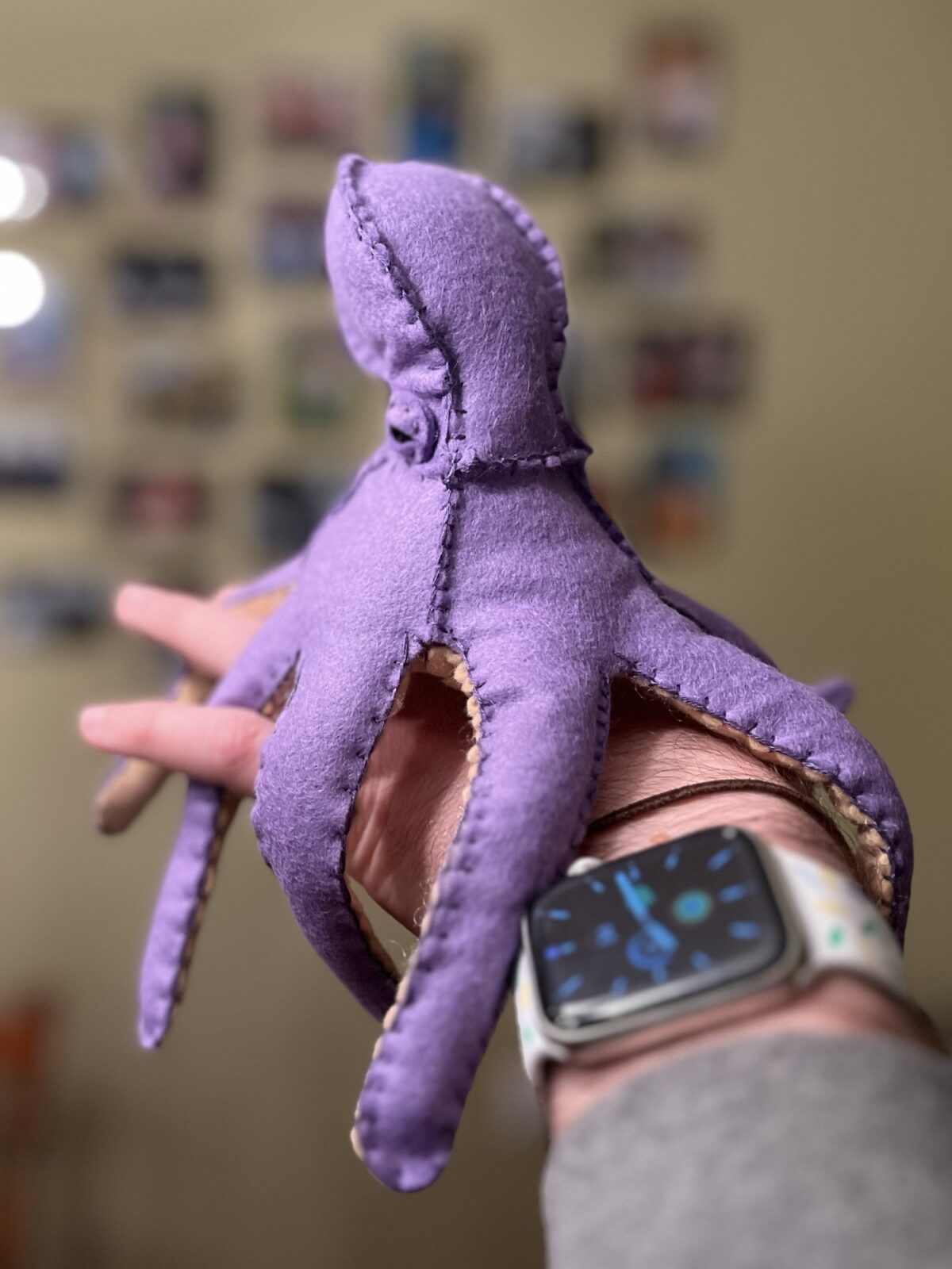 Purple felt octopus sitting on the back of my left hand with tentacles wrapped around my fingers and wrist.