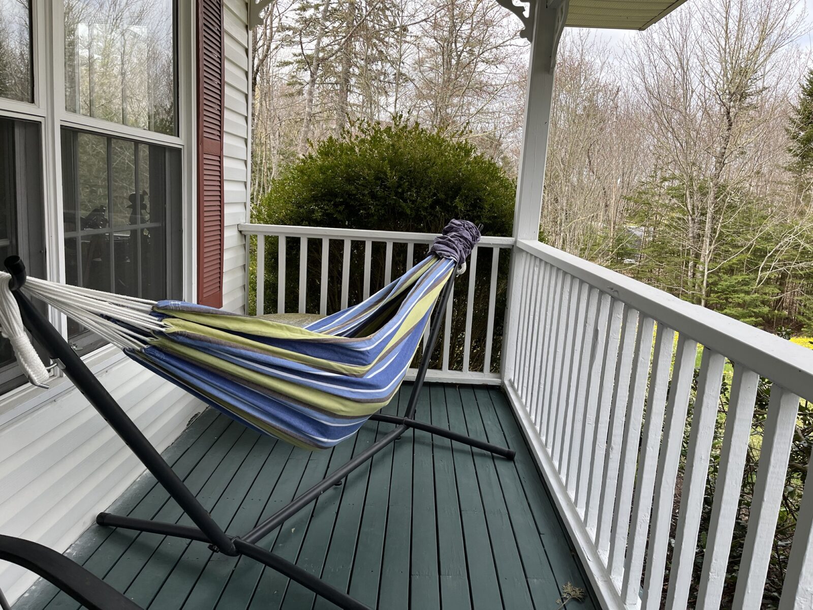 A green, blue, and white hammock hanging on a black hammock stand in the corner of our front porch