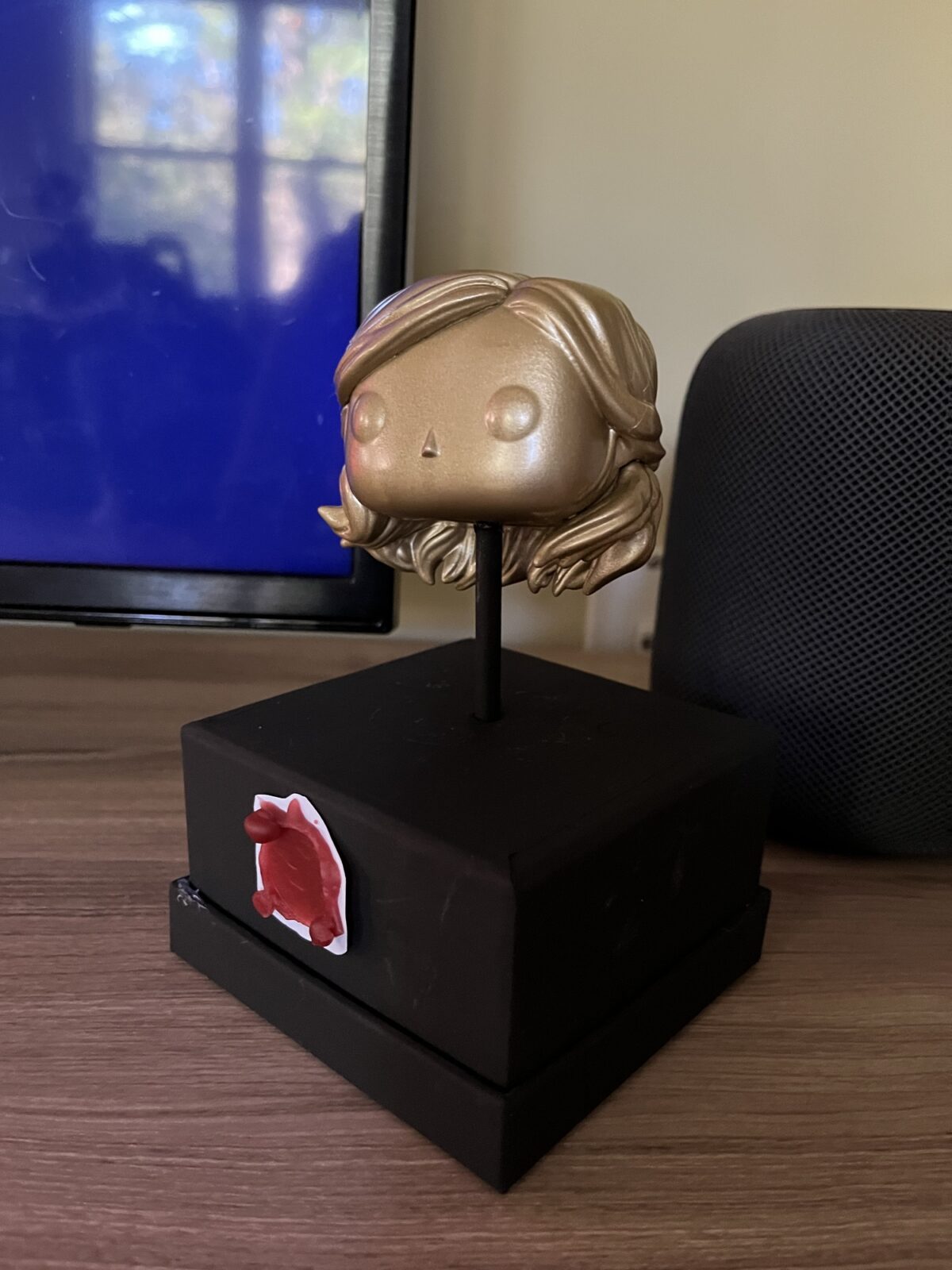 A gold, long haired, funko head mounted on a black base with a red wax seal on the base in the style of the trophy from the popular British show Taskmaster
